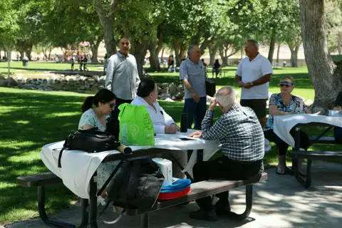 Families gathered around picnic tables under shade trees at the parish picnic