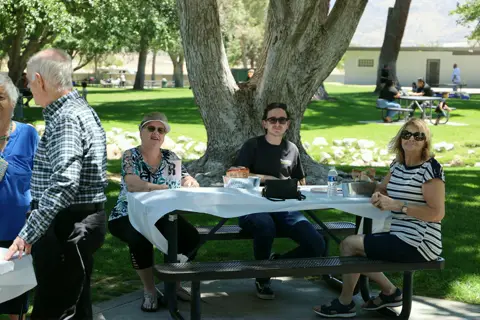 A family seated together at a long white picnic table in the shade